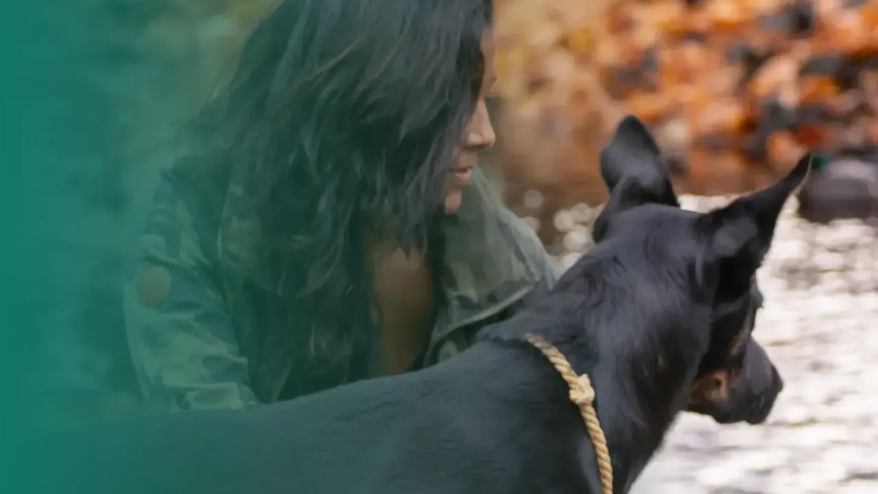 En kvinna med mörkt hår och kamouflagejacka visar sitt ledarskap hund vid en vattensamling.