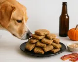 En hund nosar på en hög med hundkakor. Bredvid finns en pumpa, en flaska och lite bacon.