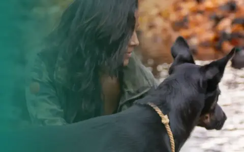 En kvinna med mörkt hår och kamouflagejacka visar sitt ledarskap hund vid en vattensamling.