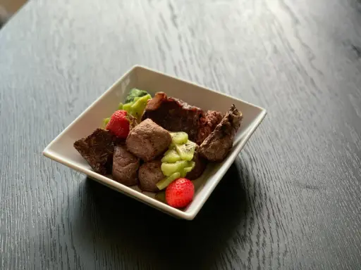 En skål med matberikning för hund: köttbitar, kiwi och jordgubbar. En näringsrik och god måltid för din fyrbenta vän.