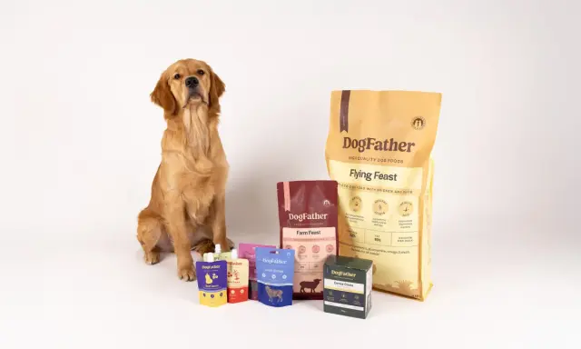 Golden retriever sitter bredvid DogFather-foder. Förpackningar med foder till hund som fiser, som "Flying Feast" och "Farm Feast", samt godis och såser.